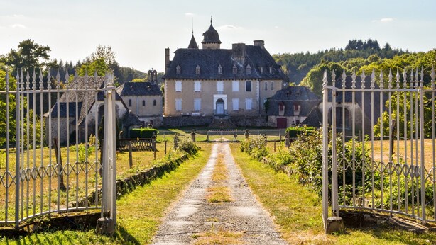 Um edifício histórico com entrada central, rodeado por um jardim formal e um caminho de cascalho que leva aos portões.