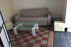 Una sala de estar con una mesa de centro de vidrio, un sofá color beige, un reposapiés blanco y baldosas de suelo con diseño.