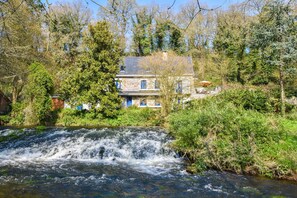 Une maison en pierre avec une cascade devant, entourée d’arbres.