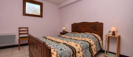 A bedroom with a wooden bed, a chair, a bedside table with a lamp, and a window with a view of trees.