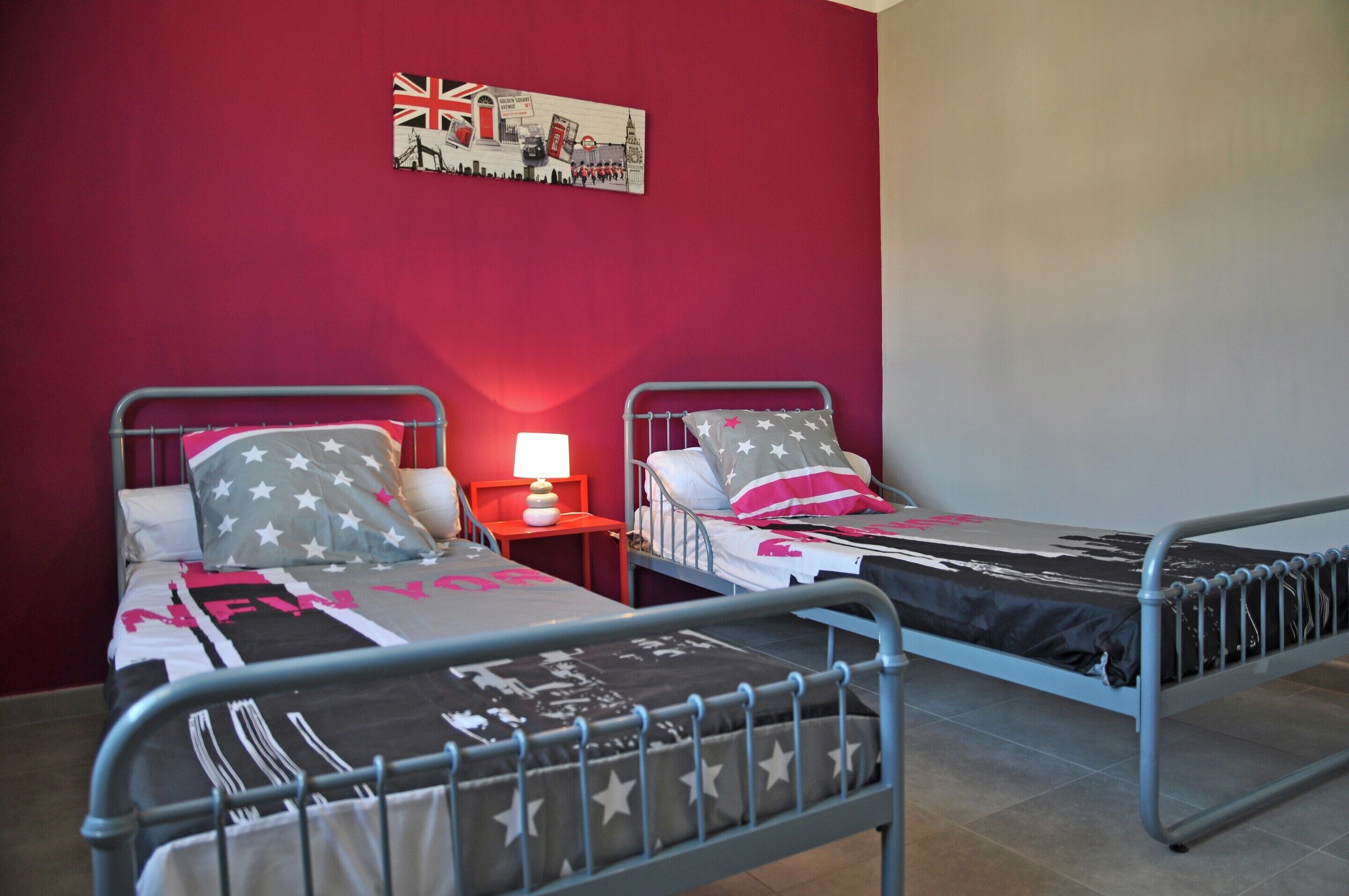 Habitació amb dos llits superposats metàl·lics, una paret vermella, una taula petita amb una làmpada i una decoració amb bandera.