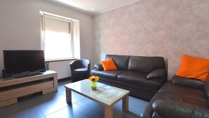 A living room with a television on a wooden stand, a coffee table with a vase of flowers, a sofa with orange cushions, and a patterned wall. - Lovely Modern Apartment, Near the Mosel, Shops and Restaurants (Zell (Mosel))