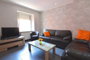 Una sala de estar con un televisor sobre un mueble de madera, una mesa de centro con un jarrón de flores, un sofá con cojines naranjas y una pared con diseño.