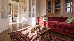 Une porte ouverte donne sur une pièce lumineuse, où l’on aperçoit un canapé rouge, un tapis à motifs et un meuble en bois.