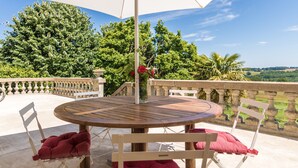 Une table ronde en bois, entourée de chaises blanches et surmontée d’un parasol blanc, est placée sur un balcon doté d’une balustrade en pierre et offrant une vue sur la verdure.
