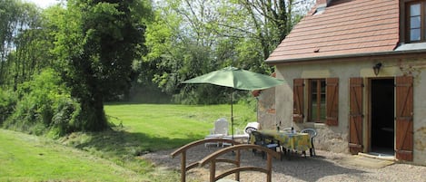 A rustic house with a gravel driveway, a wooden bridge, outdoor furniture, and a green umbrella.