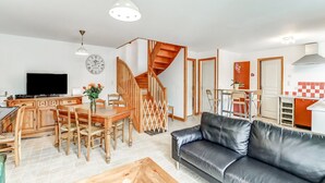 Een woonkamer met een zwarte leren bank, een houten eettafel en stoelen, een keuken met een rode en witte achterwand, en een trap.