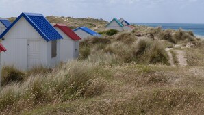 Een rij strandhutten met eenvoudige, witte muren en kleurrijke daken, gelegen op een grasachtige duin met uitzicht op zee.