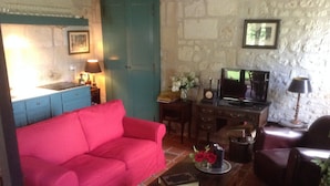 Un salon chaleureux avec un canapé rouge, une table basse en bois sur laquelle sont posés un livre et des fleurs, un téléviseur et un mur en pierre.