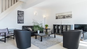 A modern living room with a sofa, coffee table, and chairs, a staircase, a flat-screen TV, and a cityscape painting. - Retreat in Beaumont-en-veron (Beaumont-en-Veron)
