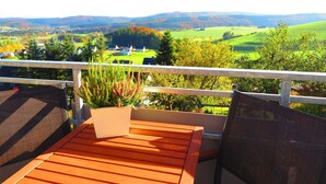 Un patio in legno con due poltrone in vimini e una pianta in vaso, affacciato su un paesaggio pittoresco con dolci colline e un campo da golf.
