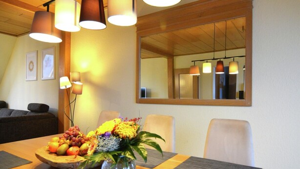Zona pranzo con tavolo in legno, ciotola di frutta e vaso di fiori. Quattro sedie attorno al tavolo, con illuminazione a sospensione visibile.