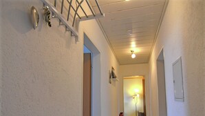 Un corridoio stretto con una porta bianca in fondo che conduce a una stanza. C'è una panca bianca con un cuscino e un oggetto rosso sopra.