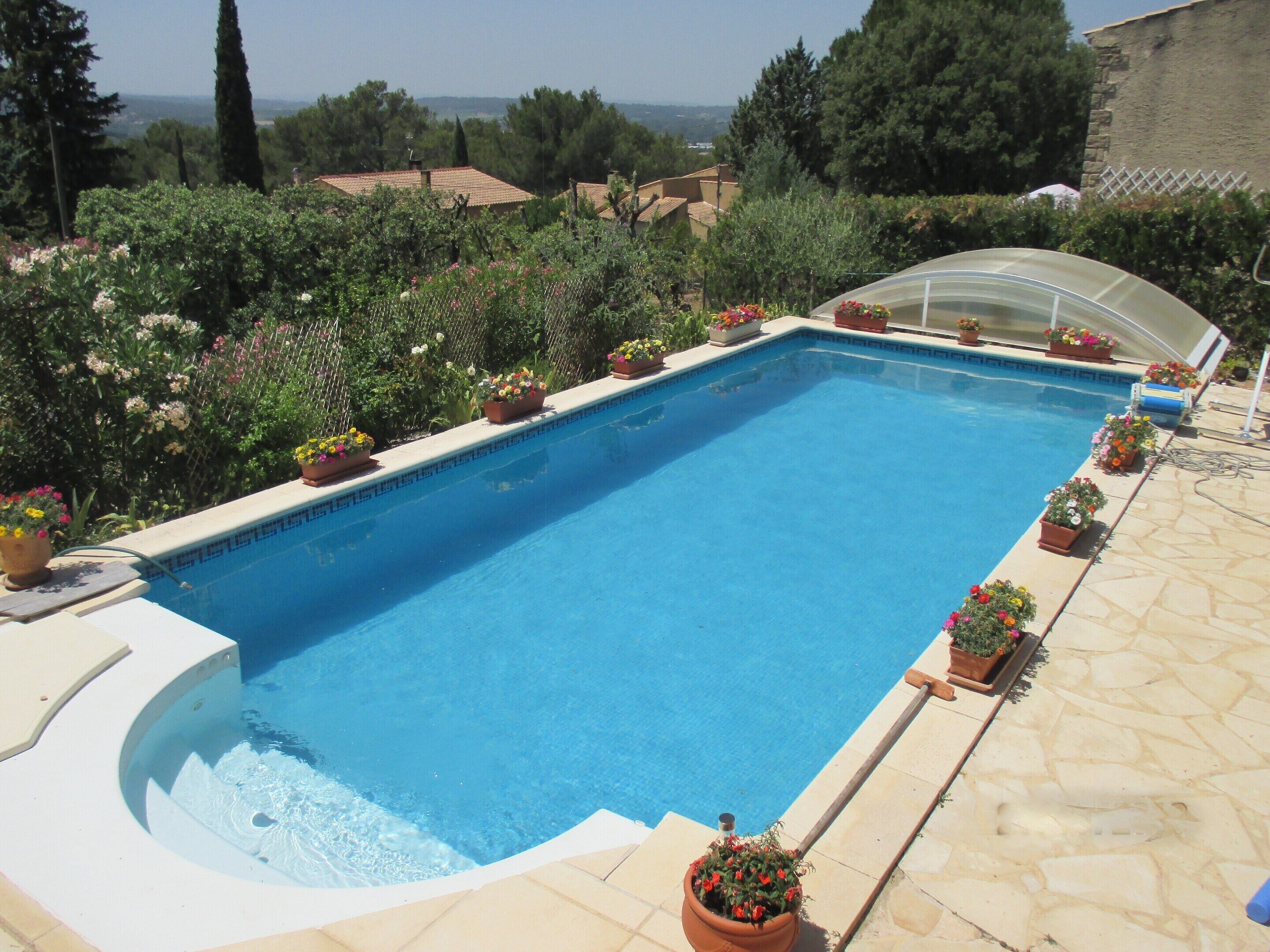 Une piscine rectangulaire dont le bord est recouvert de carreaux, entourée de plantes en pot et d’une terrasse en pierre.