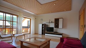Ein großzügiges Wohnzimmer mit Holzbalkendecke, einem Glastisch, einem roten Sofa, einem Fernseher auf einem weißen Schrank und einem großen Fenster mit Vorhängen.