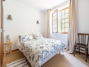 Ein Schlafzimmer mit Bett, Sessel, Nachttisch, Lampe und einem Fenster mit Vorhängen.