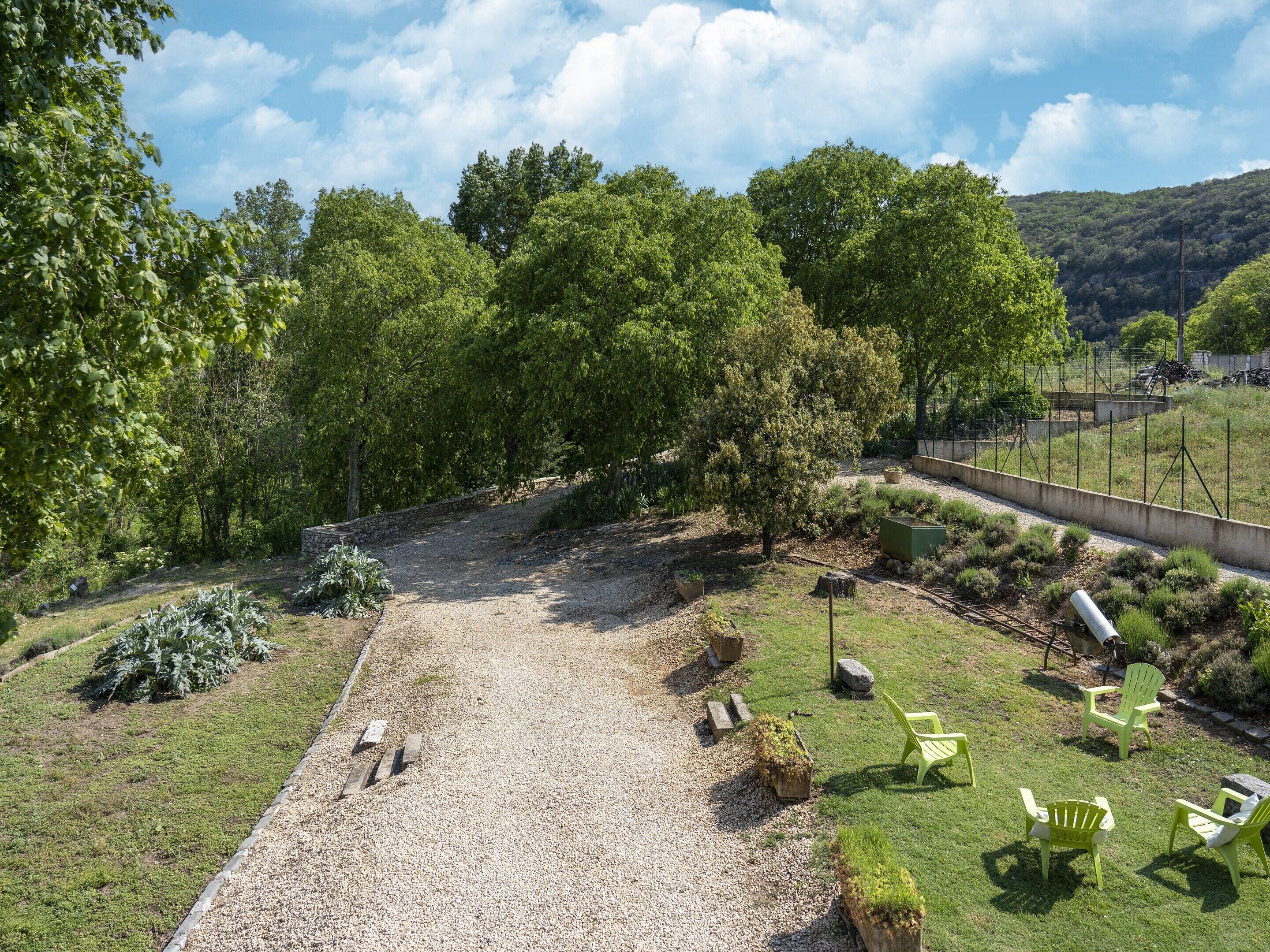 Uma trilha de cascalho serpenteia por um parque com cadeiras verdes e um muro de pedra.