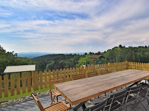 A wooden table and chairs set outdoors with a view of a forested area and a small building in the background.