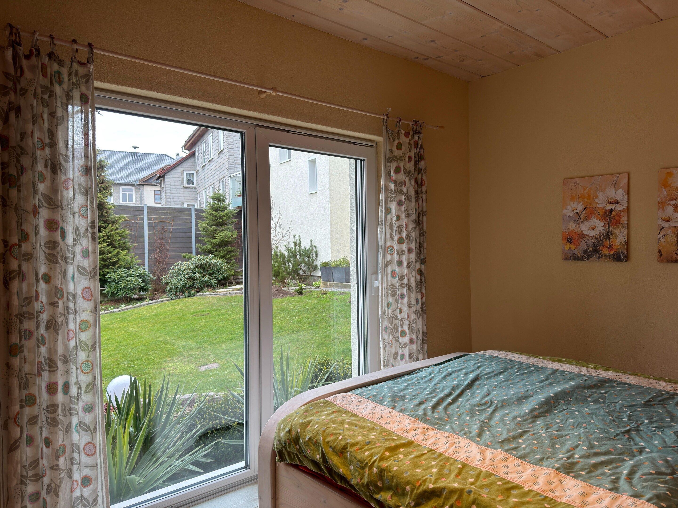 Un dormitorio con un ventanal grande, una cama con cubrecama verde y melocotón, y dos cuadros colgados en la pared.
