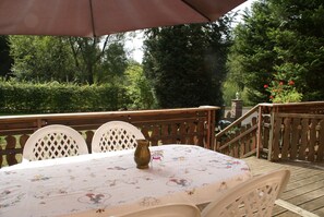 An outdoor patio with a table covered in a floral tablecloth, surrounded by white chairs, and a wooden deck with a railing.