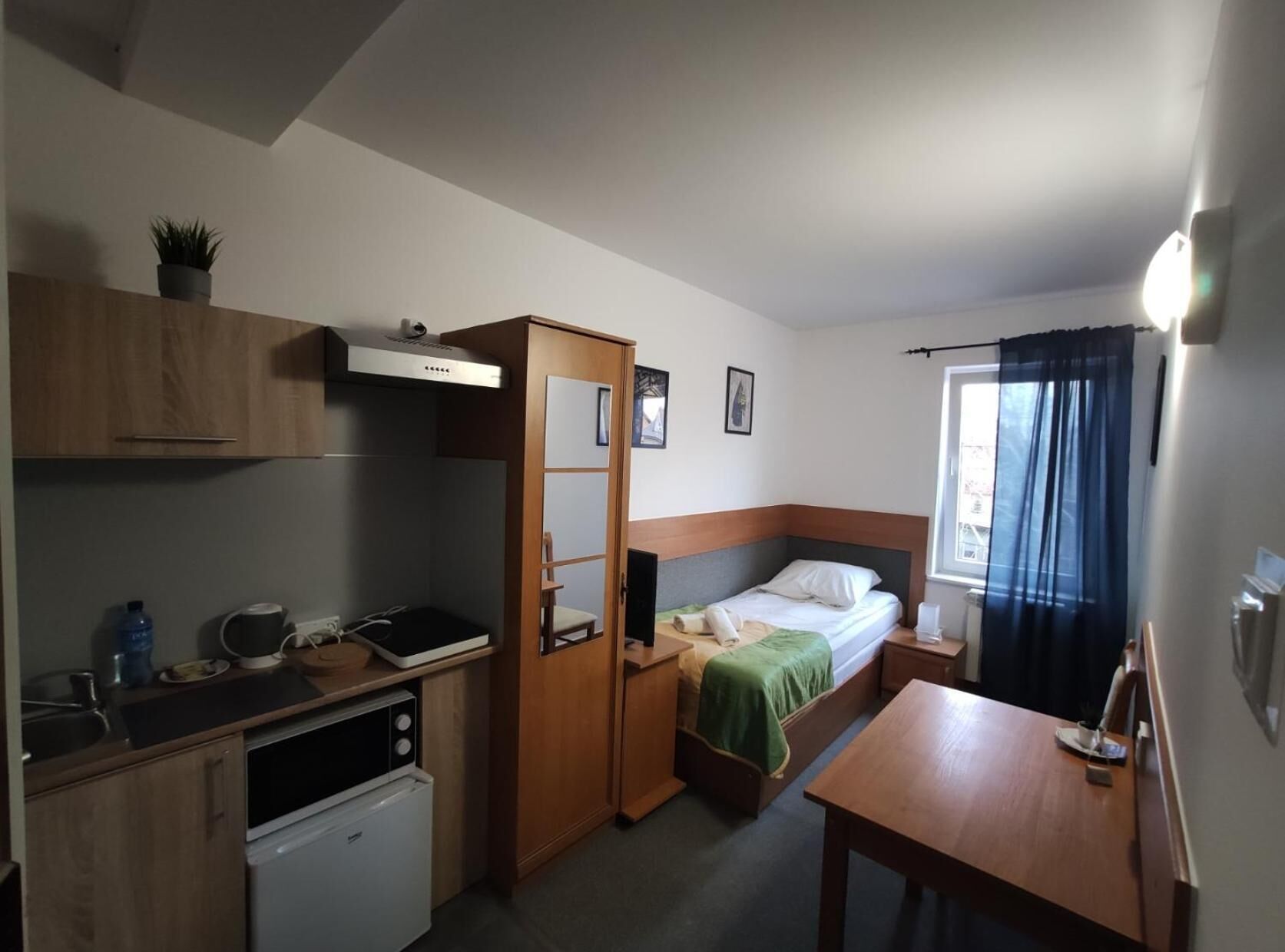 Et kompakt værelse med tekøkken, en seng med et grønt tæppe, et lille bord og et vindue med gardiner.