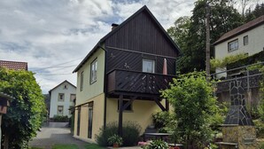 Une maison à deux étages avec un balcon en bois sombre, une allée pavée et un mur de pierre agrémenté de plantes en pot.