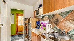 Une cuisine compacte équipée d’un micro-ondes, d’une bouilloire et d’une machine à café sur le plan de travail, d’un évier et d’une porte donnant sur un couloir avec un mur vert et une porte en bois.