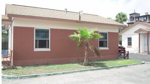 A single-story brick house with a flat roof, two windows, and a small palm tree in the front yard. - Bayfront Cottage Room 11 (Rockport)