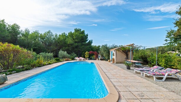 Ein rechteckiger Swimmingpool mit Fliesenrand, umgeben von einer Steindeck. Liegestühle mit Kissen und ein kleines Gebäude mit einem gefliester Dach.