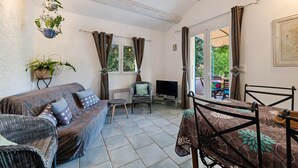 Ein gemütliches Wohnzimmer mit Sofa, Fernseher, Esstisch und Stuhl. Es gibt Fenster mit Vorhängen, eine hängende Pflanze und ein gerahmtes Bild an der Wand.
