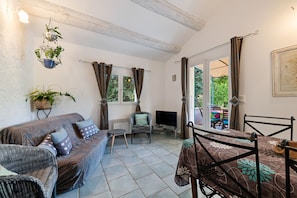 A cozy living room with a sofa, a television, a dining table, and a chair. There are windows with curtains, a hanging plant, and a framed picture on the wall. - Modern Holiday Home With Private Pool (Lambesc)