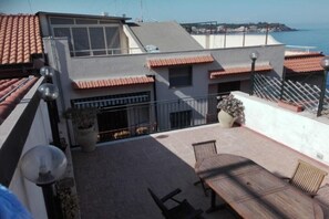 A rooftop terrace with a wooden table and chairs, a potted plant, and a view of the sea.
