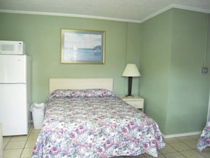 A bedroom with a bed, a nightstand with a lamp, a microwave, and a trash bin. - Bayfront Cottage Room 12 (Rockport)