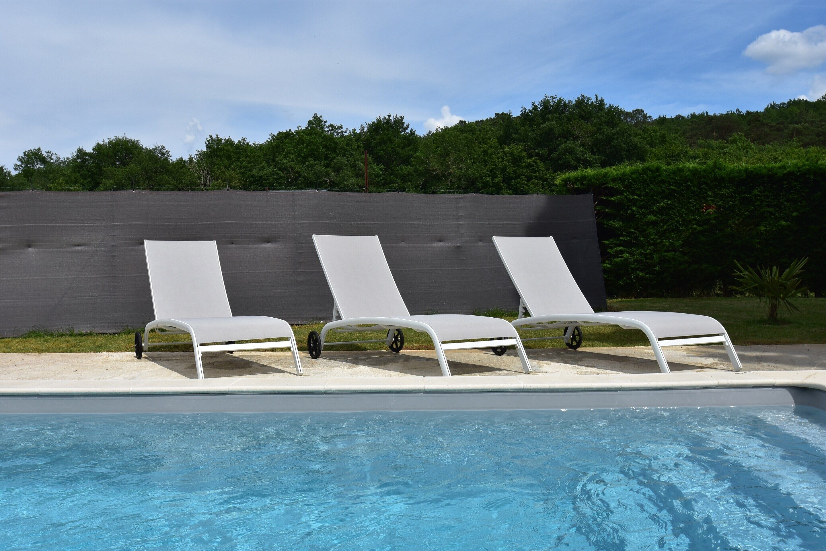 A swimming pool with three white lounge chairs and a dark wall in the background.