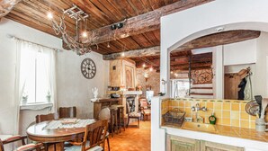Un intérieur rustique avec une table à manger en bois, des chaises, un lustre, un coin cuisine avec un dosseret carrelé et un mur en briques.