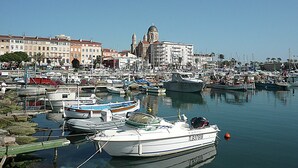 Un port de plaisance avec des bateaux amarrés, un bâtiment imposant surmonté d’un dôme en arrière-plan et un ciel bleu dégagé.