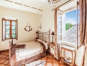 Ein Schlafzimmer mit Bett, einem Fenster mit Vorhängen, einem Nachttisch mit Lampe und einem kleinen Tisch mit Radio.
