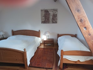 Quarto com duas camas de madeira, uma mesinha de cabeceira com abajur, tapete vermelho estampado e um vigamento de madeira no teto.