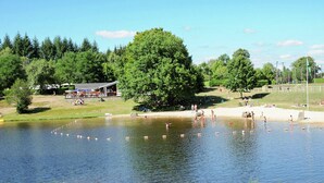 A lakeside setting with a large tree, a building with a roof, and a sandy area with people. - Beautifully Located Country House With Heated Pool (Auriac)