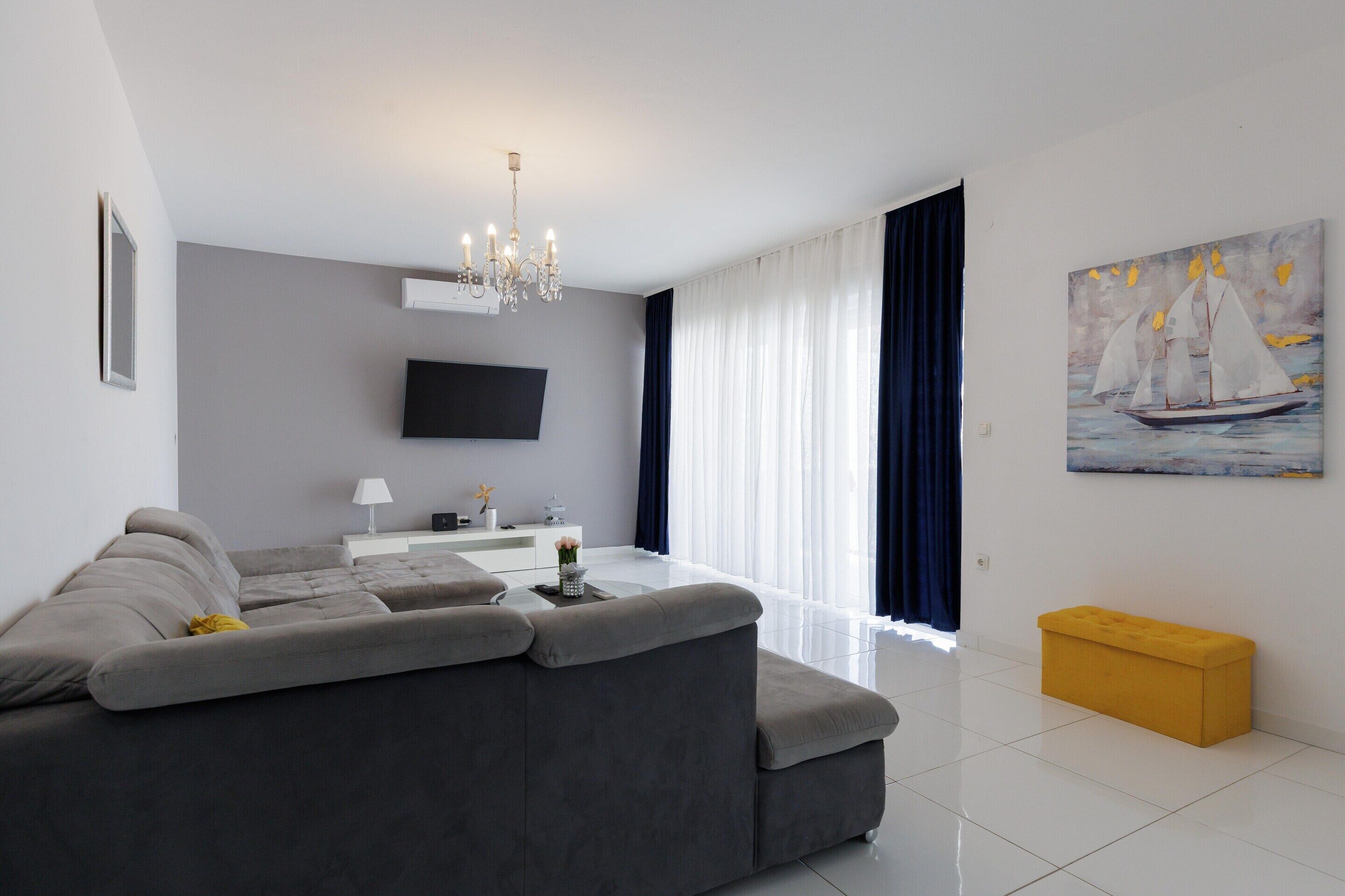 Un salón moderno con un sofá gris, un televisor de pantalla plana, una araña y un cuadro de veleros.