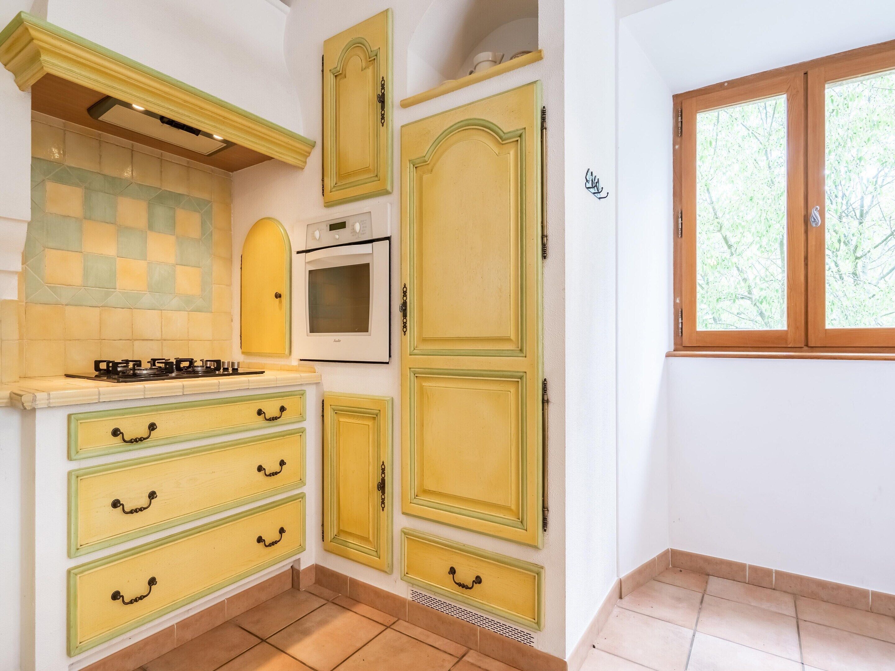 Une cuisine avec des meubles de couleur jaune, une cuisinière, un four et une fenêtre donnant sur des arbres.