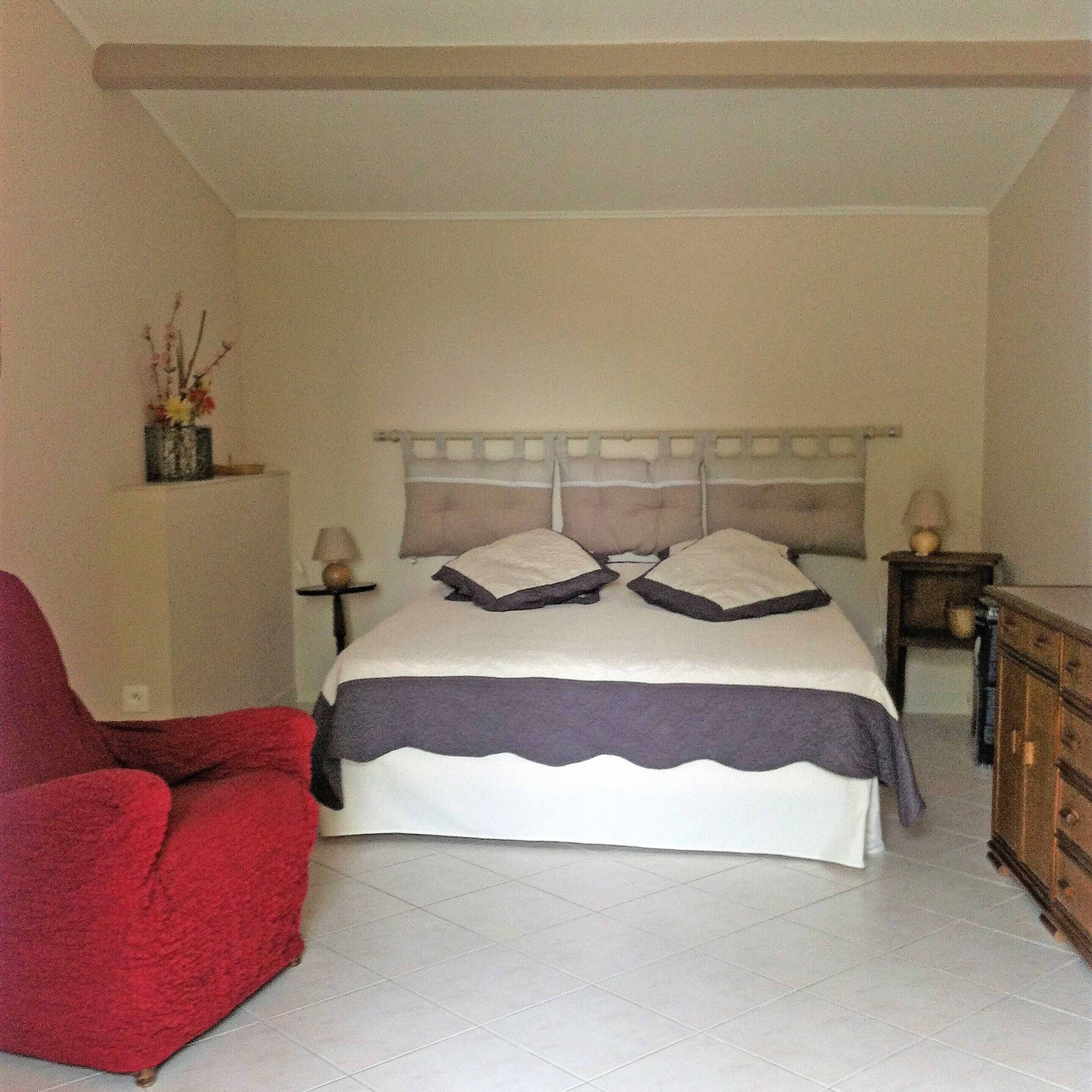 A bedroom with a bed, a red chair, a dresser, and a nightstand with a lamp.