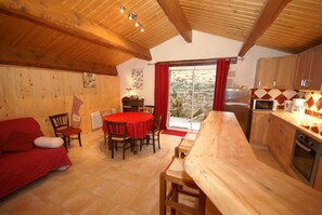 A cozy room with a wooden ceiling, a dining table with chairs, a red sofa, and a kitchen area with cabinets and a stove. - House in Ardeche Near Ray-pic Falls (Saint-Pierre-de-Colombier)
