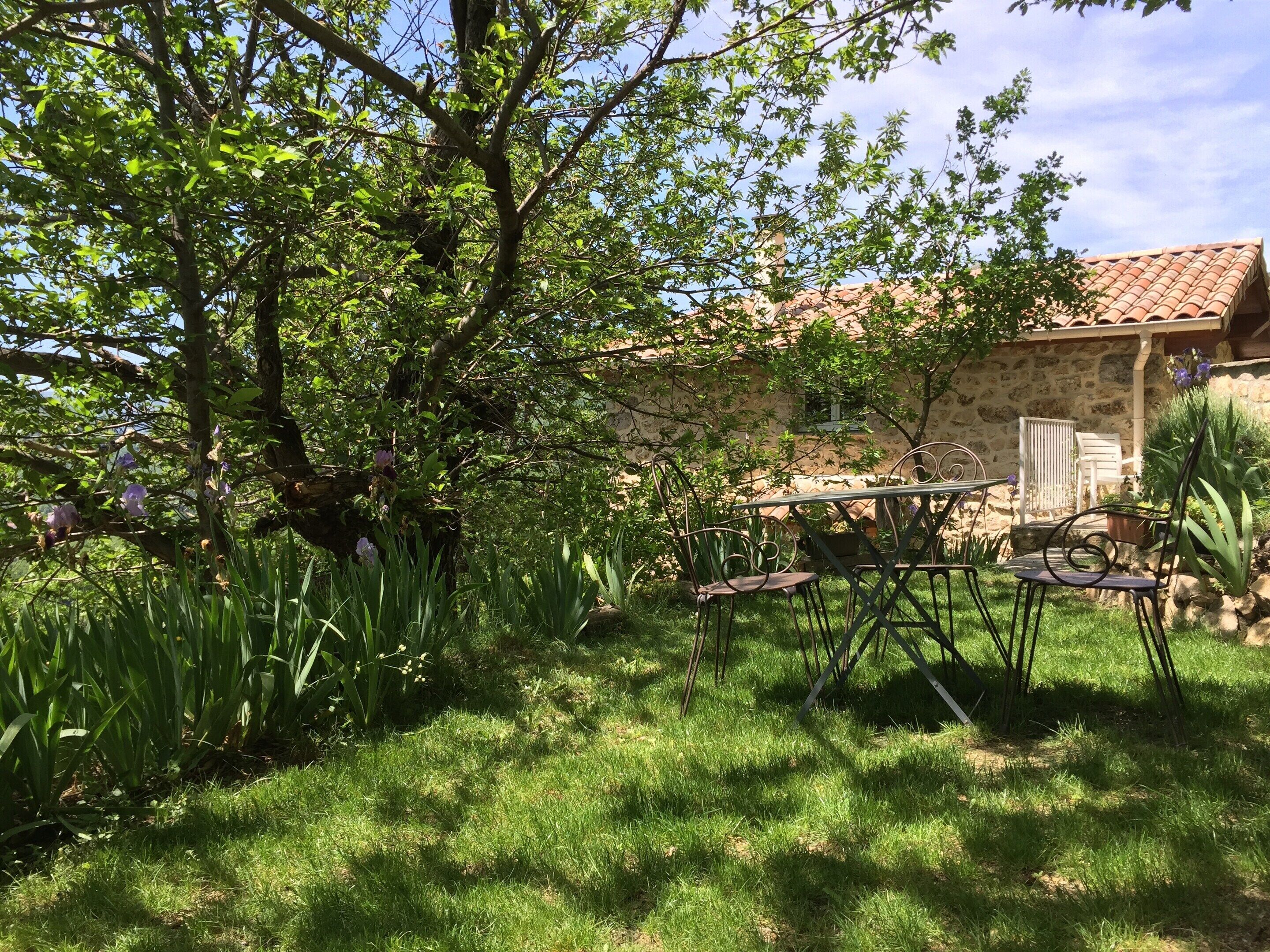 A stone building with a tiled roof, surrounded by lush greenery and a grassy area with metal chairs.