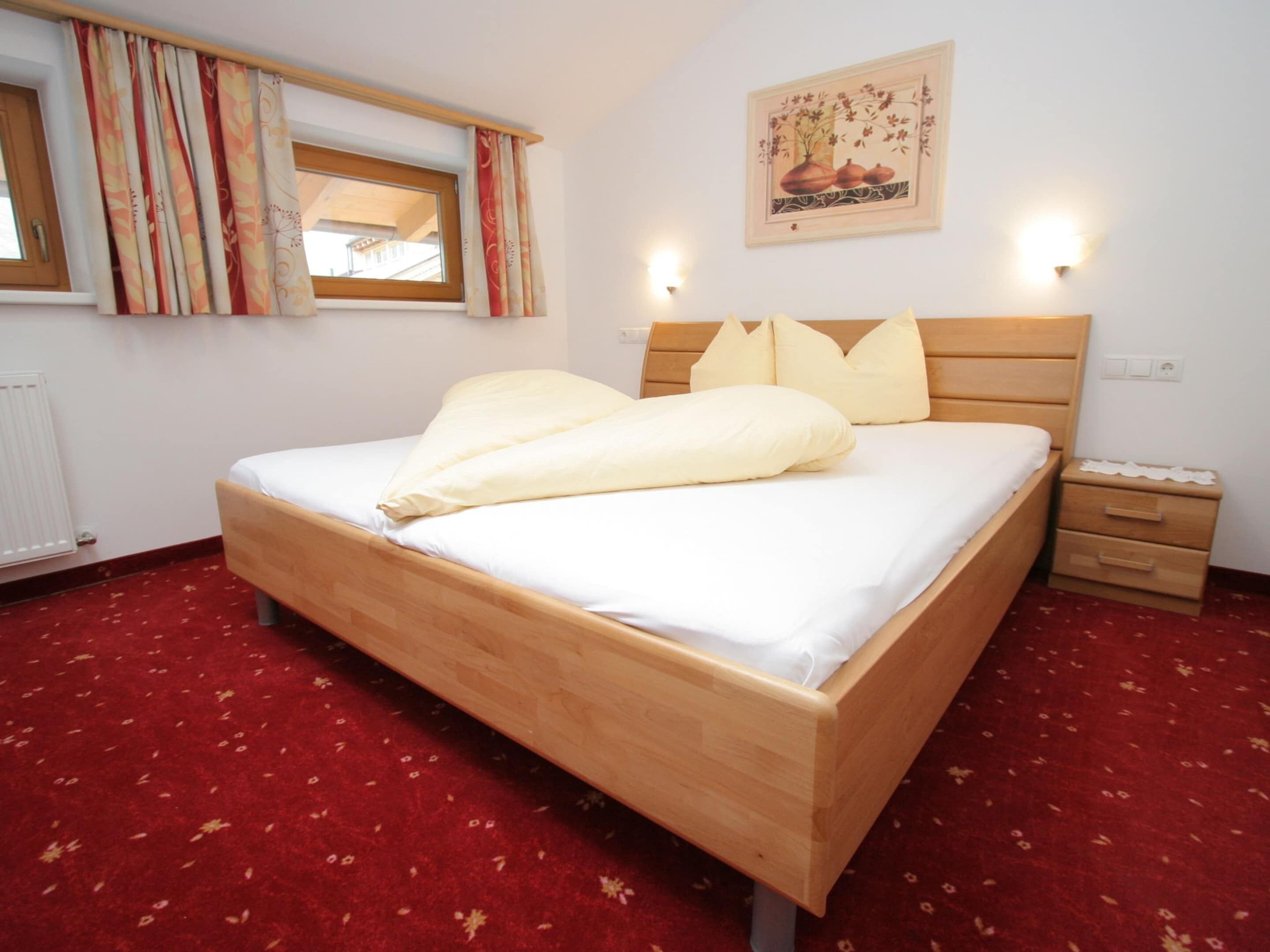 Un lit bien fait, avec du linge de lit blanc et deux oreillers, une tête de lit en bois, une table de chevet avec des tiroirs, une fenêtre avec des rideaux et un luminaire fixé au mur.
