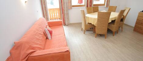 A room with a sofa, a dining table with chairs, a wooden bench, and a window with curtains.