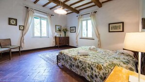 Un dormitorio con una cama, una silla, un aparador de madera, una lámpara y una ventana con cortinas.