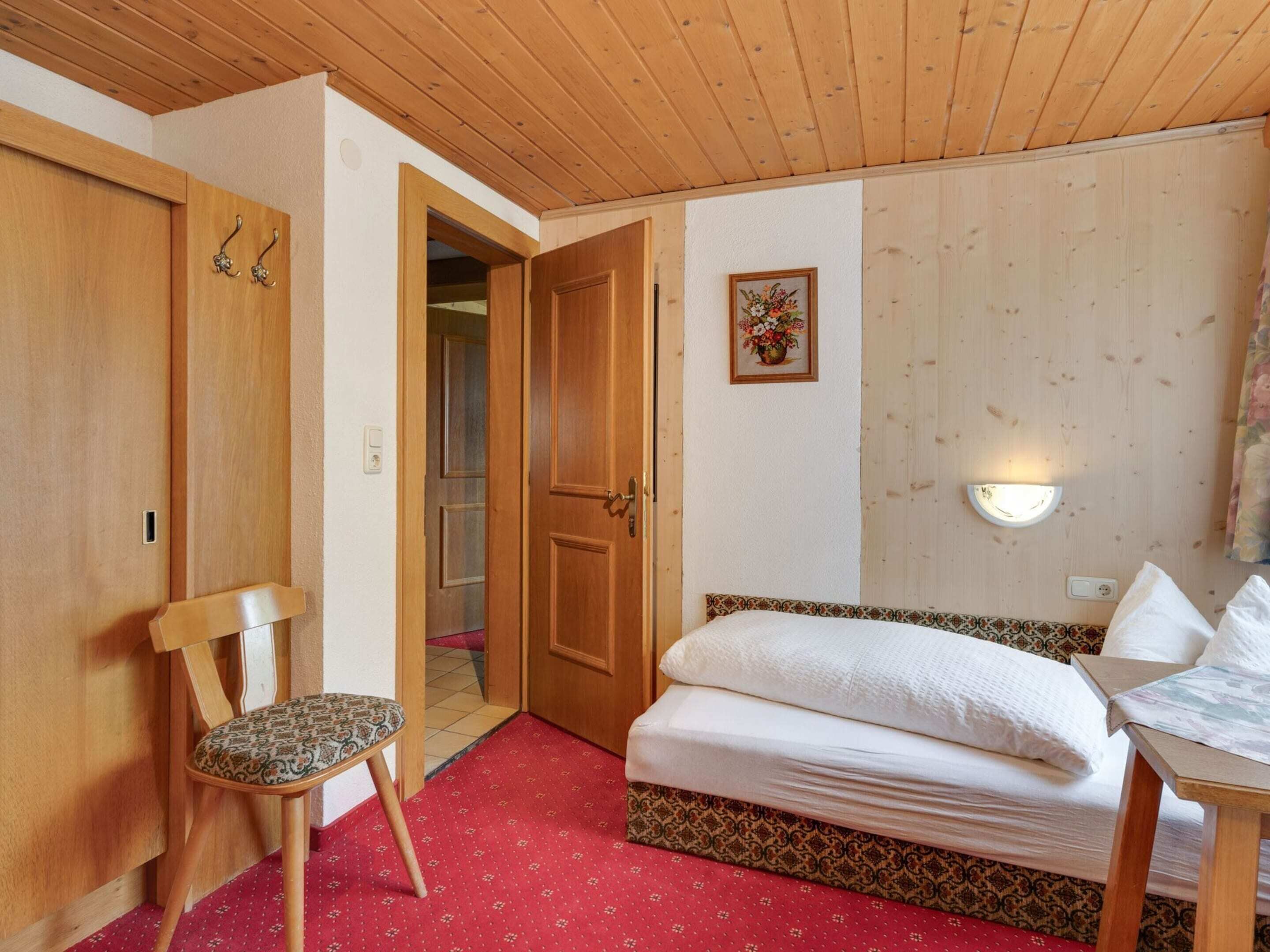Une chambre avec un plafond en bois, un lit recouvert de linge de lit blanc, une chaise en bois, un tapis rouge à motifs et un luminaire fixé au mur.