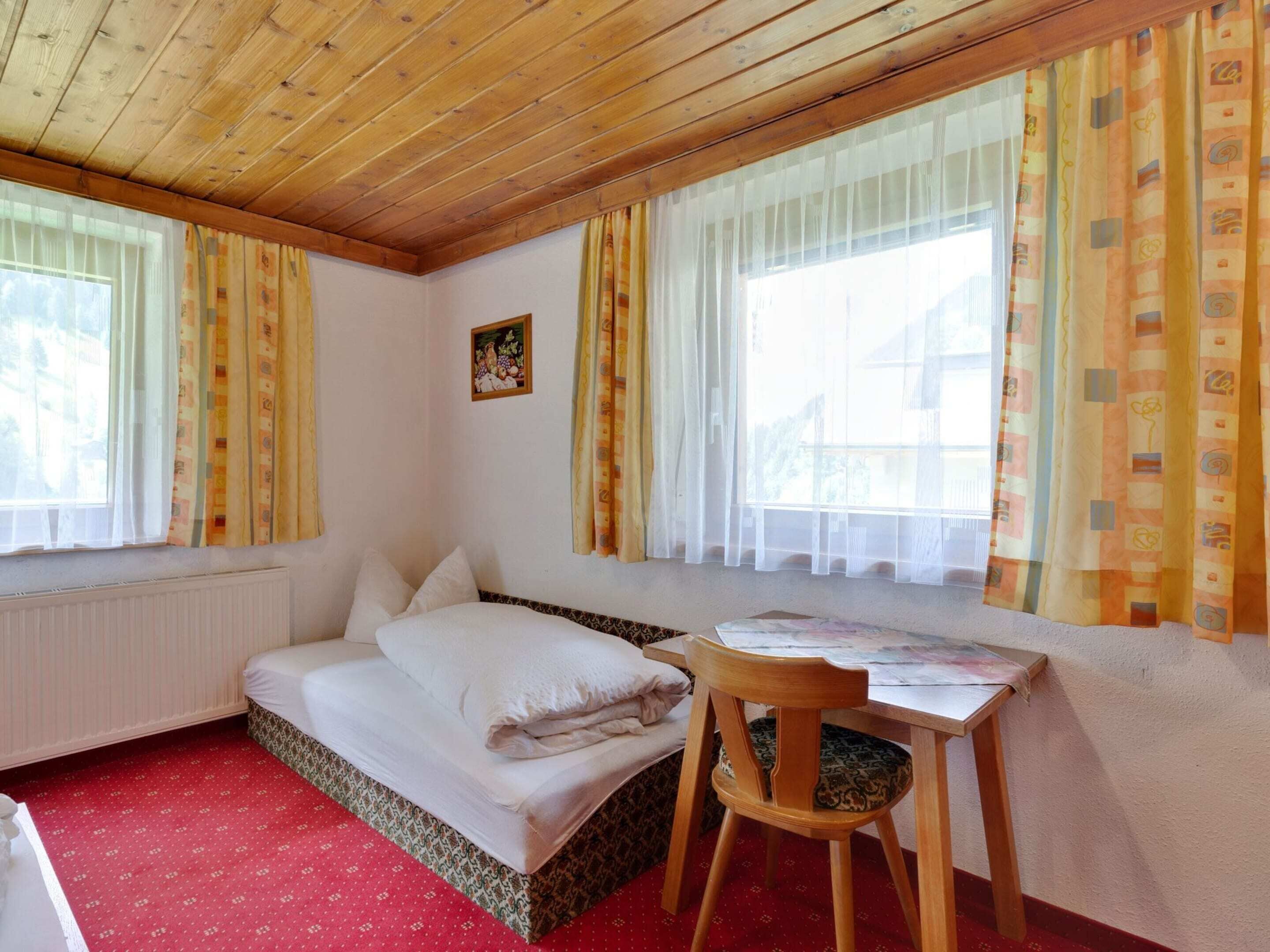 A cozy room with a bed, a wooden chair, a table, and windows with curtains.