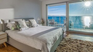 A modern bedroom with a large bed, a nightstand, a lamp, and a view of the sea through a glass door. - Luxurious Villa in Kozino With Fenced Garden (Zadar)